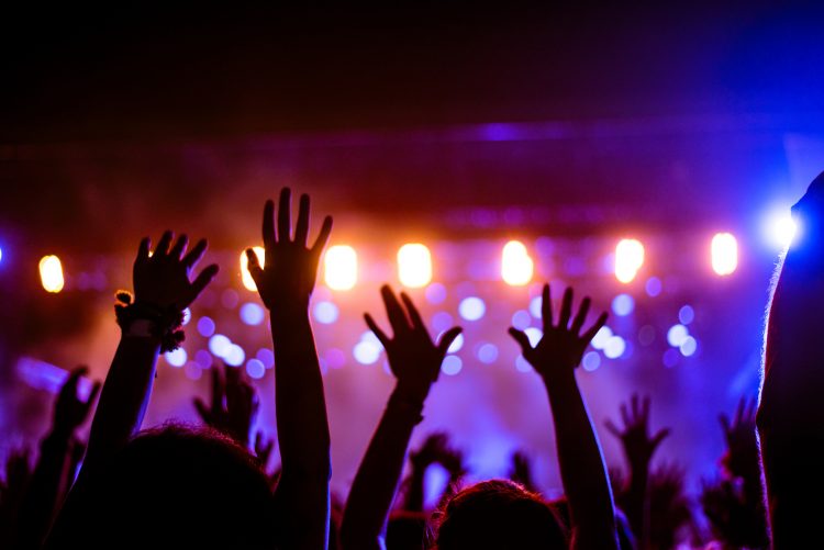 Audience with hands raised at a music festival and lights streaming down from above the stage. Soft focus, blurred movement.