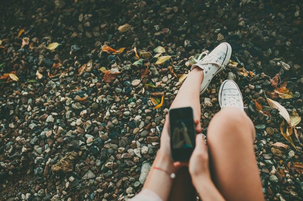 Young hipster girl making a photo of legs with a smartphone on rocky coast background. Feet selfie with copy space.